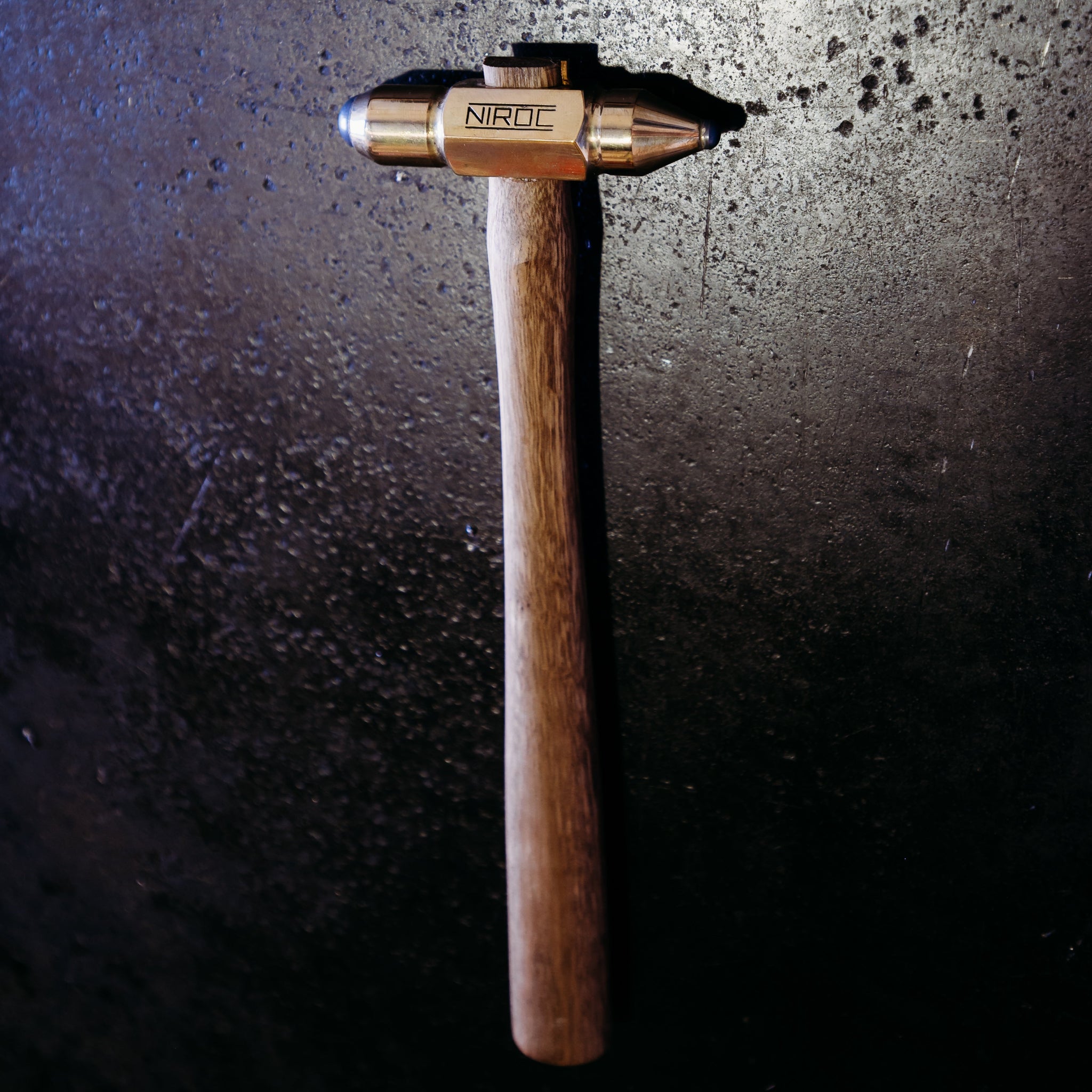 A Niroc brand hammer with a wooden handle and brass-colored head, photographed against a dark, textured background.
