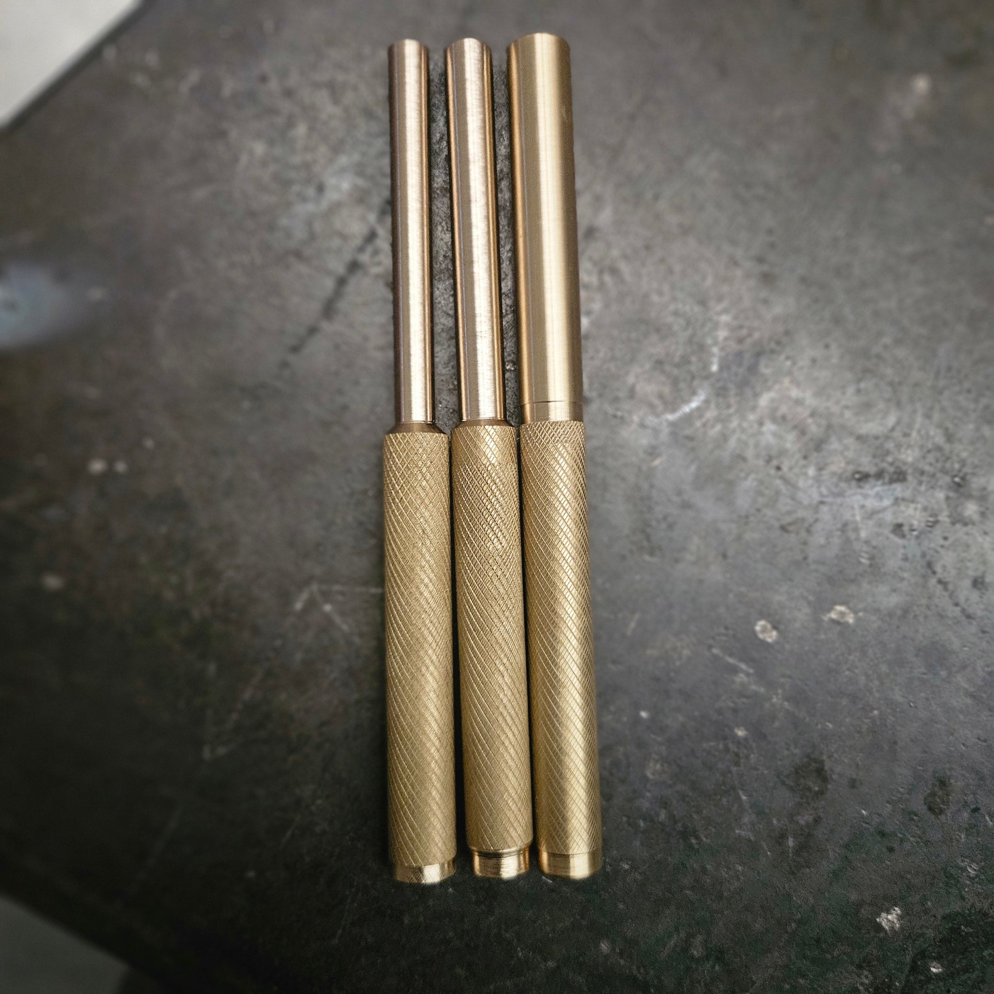 Three brass drifts with knurled textured handles and smooth cylindrical shafts, arranged vertically on a dark, scratched surface.